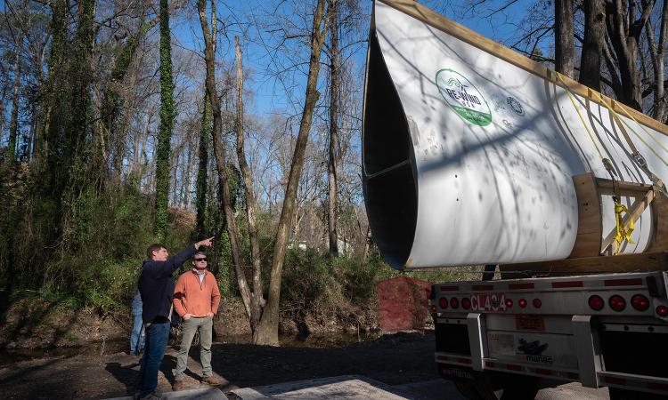 Two people standing in a wooded area point and discuss a large wind turbine blade sitting on a truck trailer. (Photo: Allison Carter)